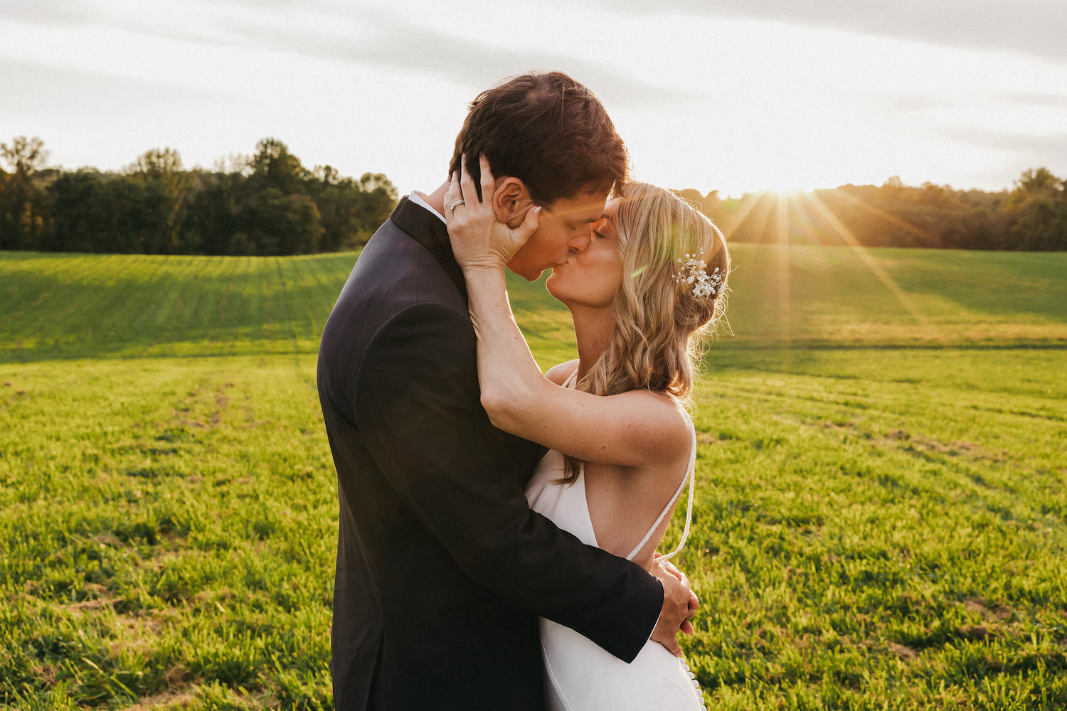 Bride and groom sharing a kiss in the middle of a vast green field with the sun setting in the horizon behind them, captured by Rachel Yearick Photography