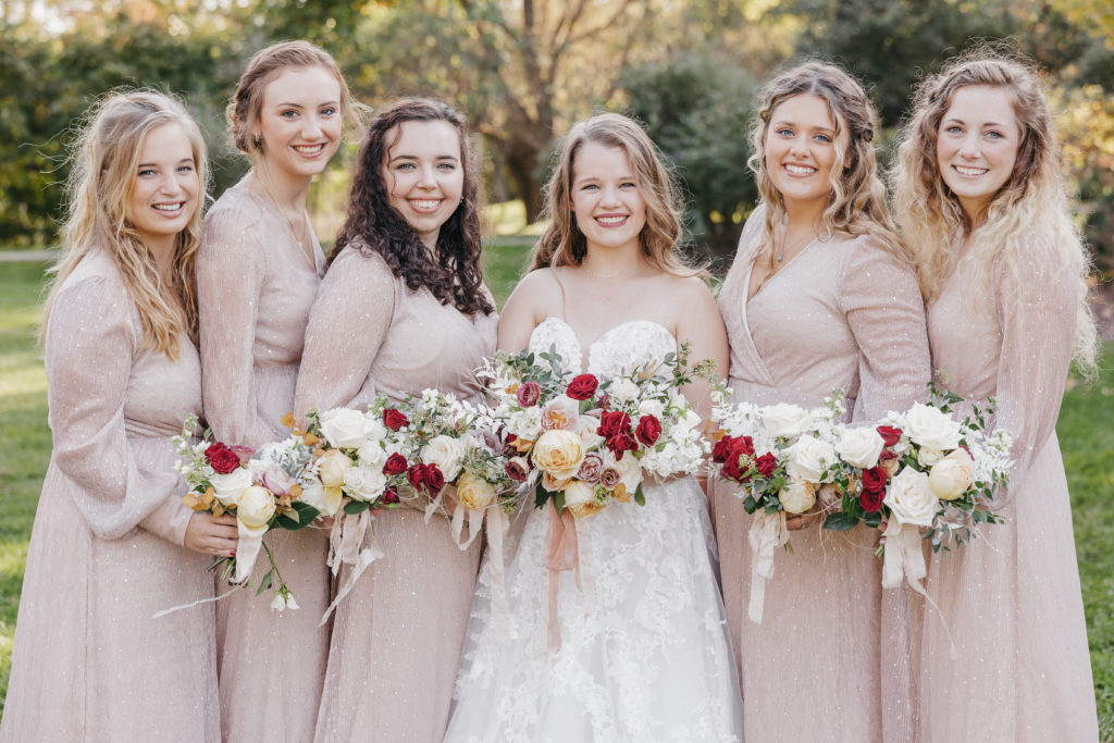 Close-up shot of bride posing with her bridesmaids in a line as they hold up their white rose bouquets, taken by Rachel Yearick Photography