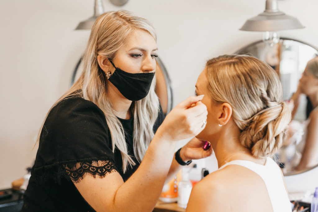 Ultimate Wedding Vendor List (Checklist Included). Bride getting her makeup done by a makeup artist with a mask on.