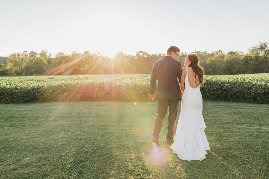 Bride and groom holding hands and smiling at each other as sun shines in front of them, taken by Rachel Yearick Photography