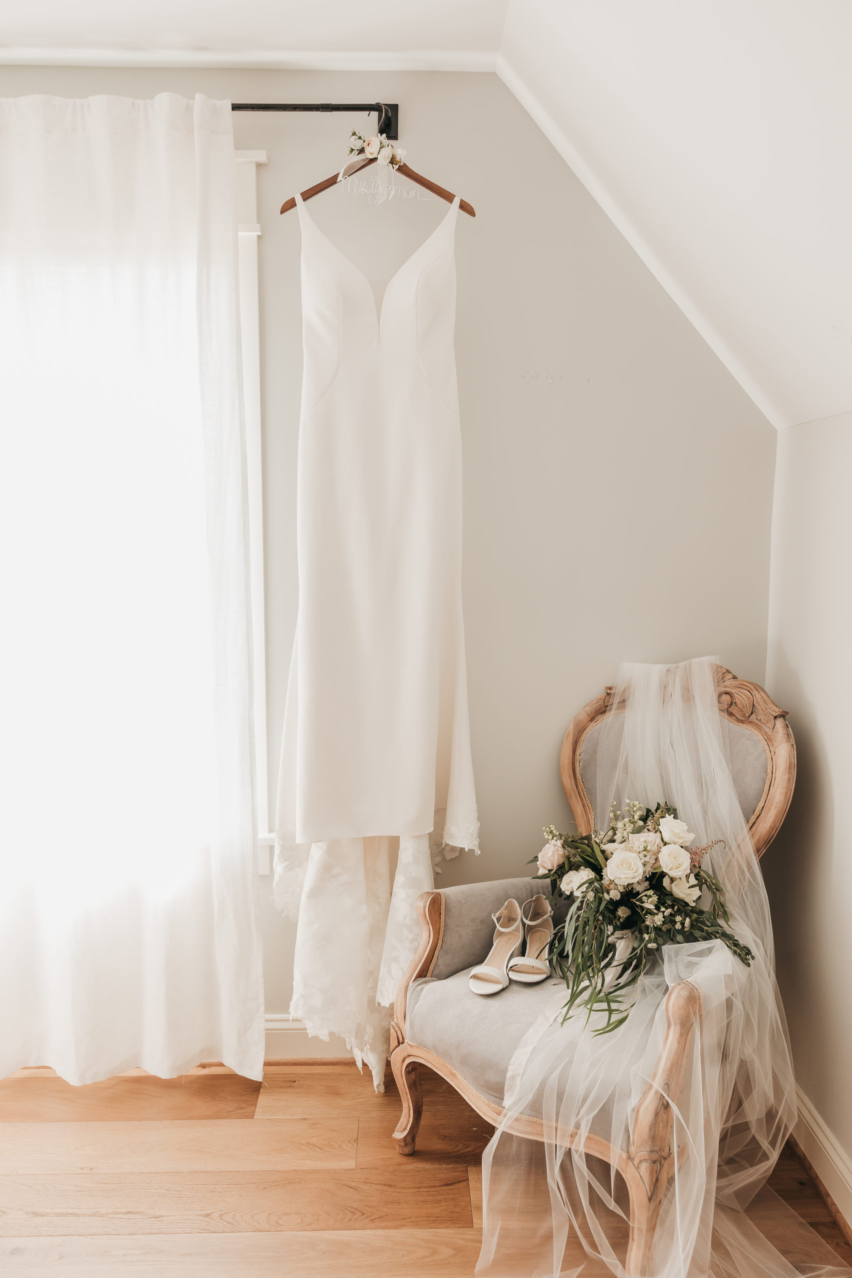Bride's dress hung from curtain rod with veil, bouquet and shoes on chair beside it, taken by Rachel Yearick Photography