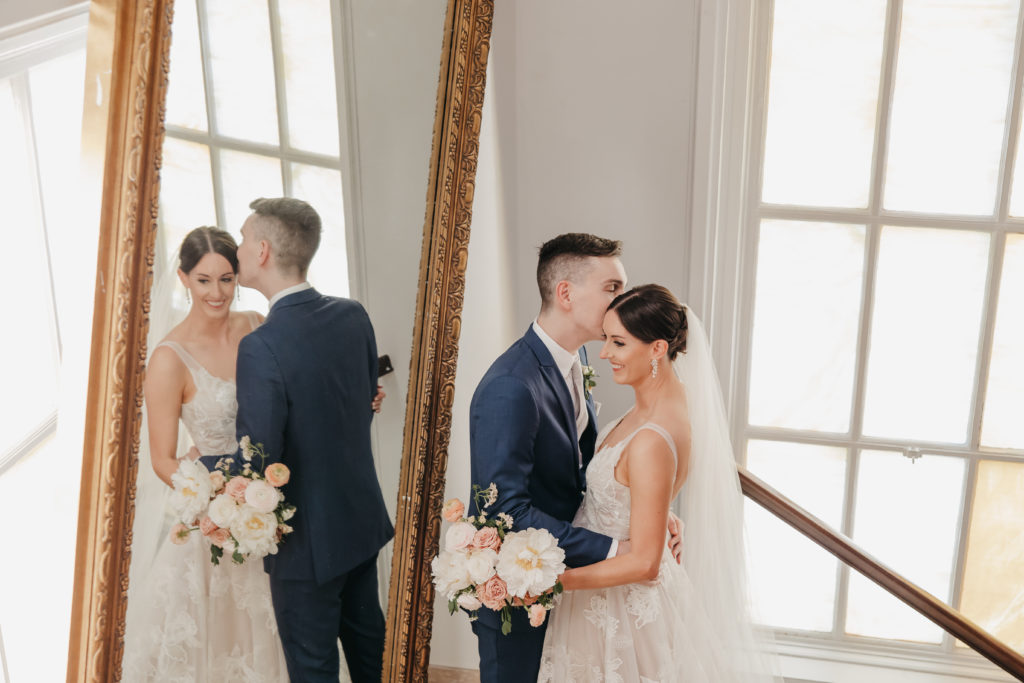 Groom planting a kiss on bride's forehead as she smiles with mirror beside them, shot by natural light wedding photographer Rachel Yearick