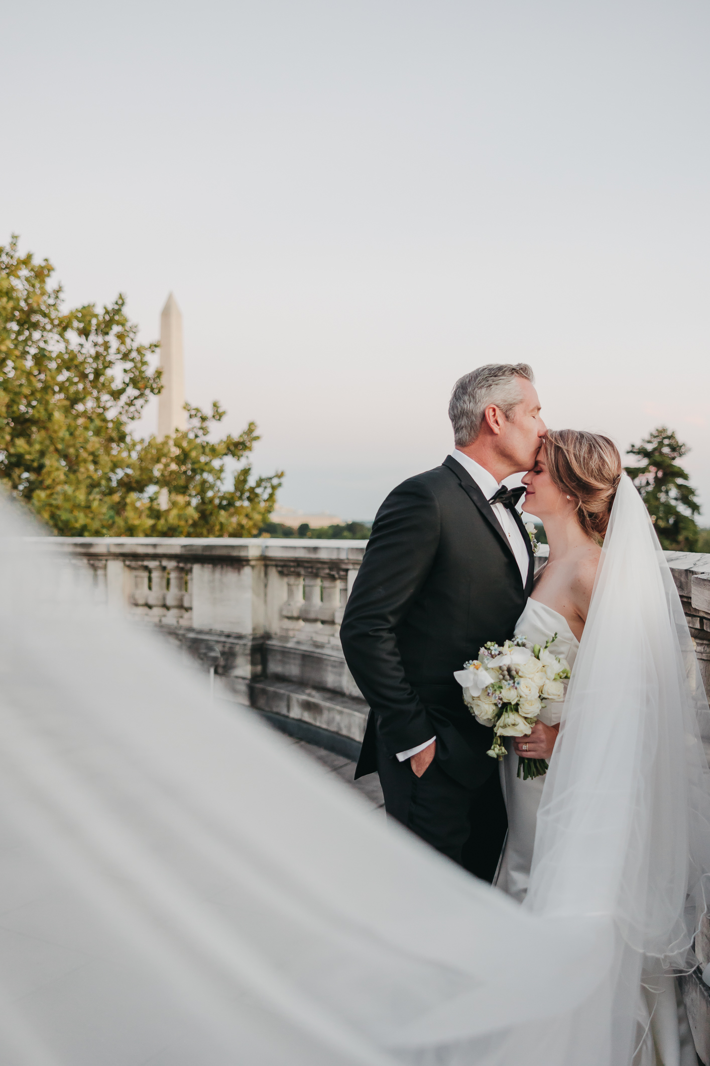 Groom plants a kiss on bride's forehead as her veil sways in the wind, taken by Rachel Yearick Photography