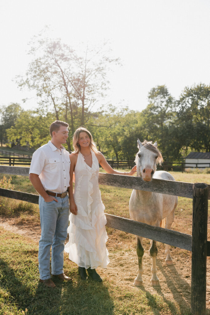 Bride-to-be feeding a horse while her fiancé smiles beside her during a relaxed countryside engagement session.