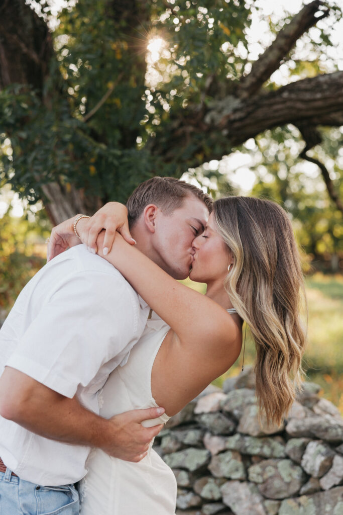 Golden-hour portrait of an engaged couple kissing in an open meadow, surrounded by charm natural light