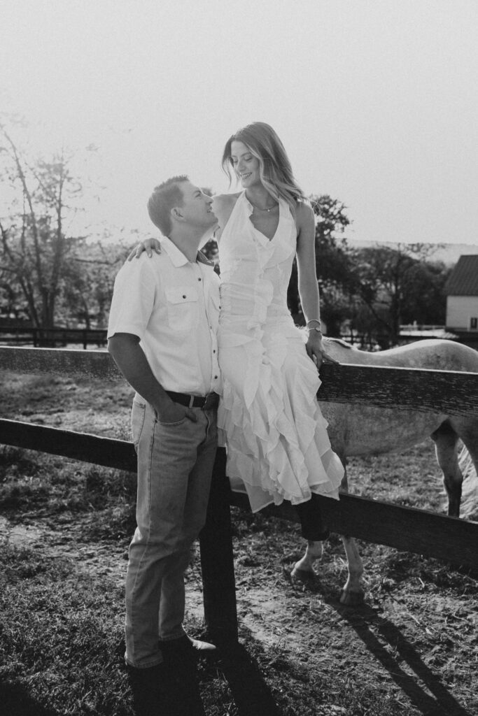 Romantic countryside engagement photo of Olivia and Andrew in Bluemont, Virginia, standing by a wooden fence with a horse under soft golden light.