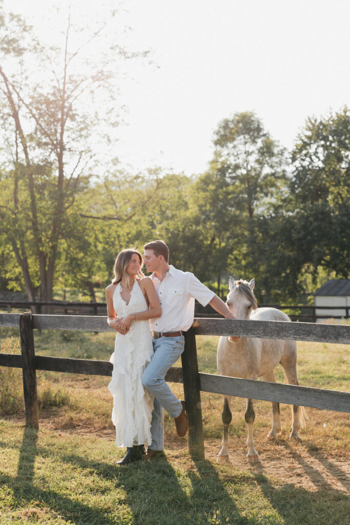 Romantic countryside engagement photo of Olivia and Andrew in Bluemont, Virginia, standing by a wooden fence with a horse under soft golden light.