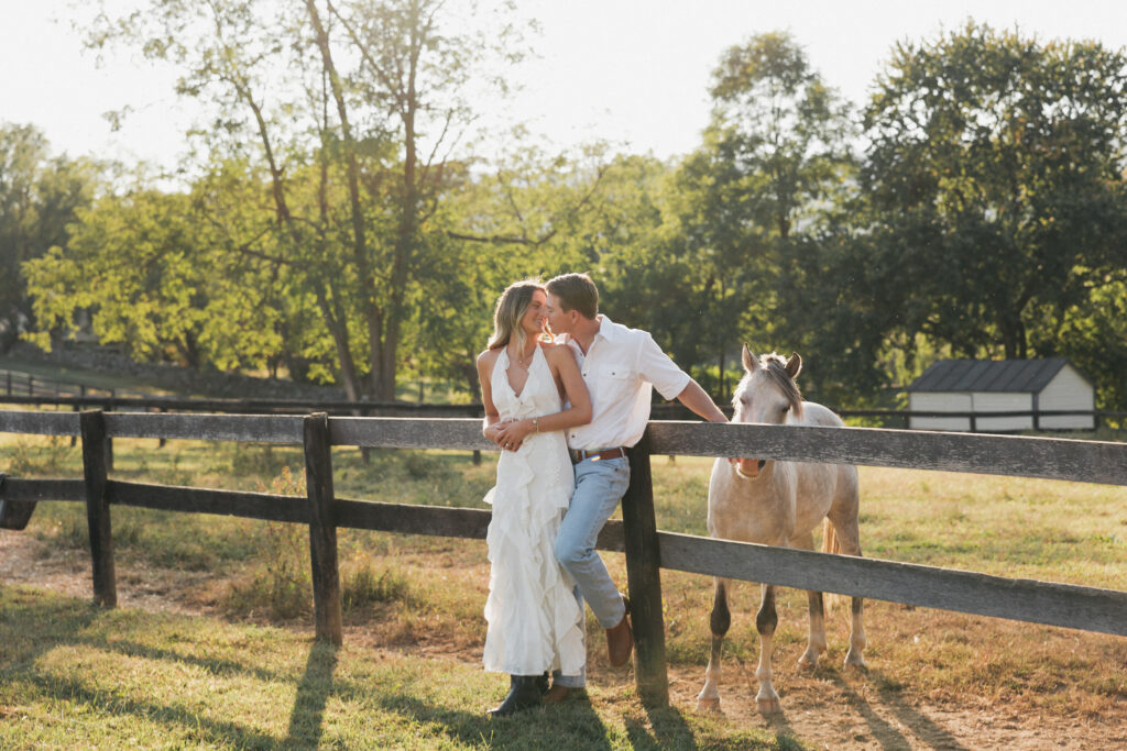 Romantic countryside engagement photo of Olivia and Andrew in Bluemont, Virginia, standing by a wooden fence with a horse under soft golden light.